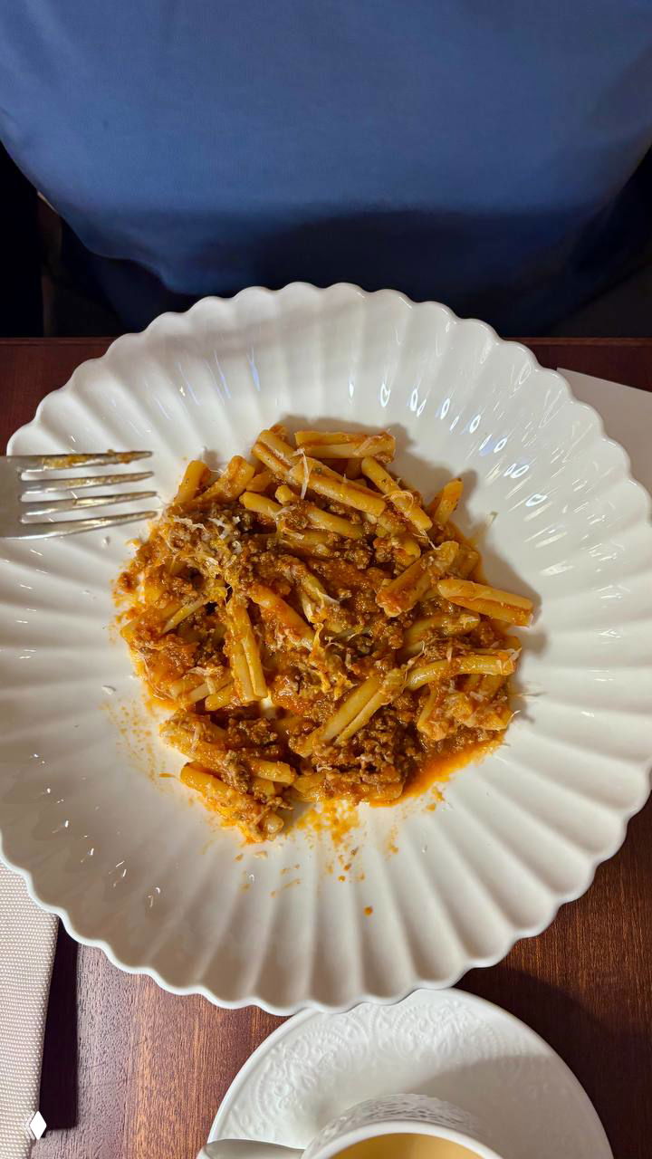 Паста с мясным соусом (рагу) в белой волнистой тарелке: плотная текстура, насыщенный томатный соус и тертый сыр.