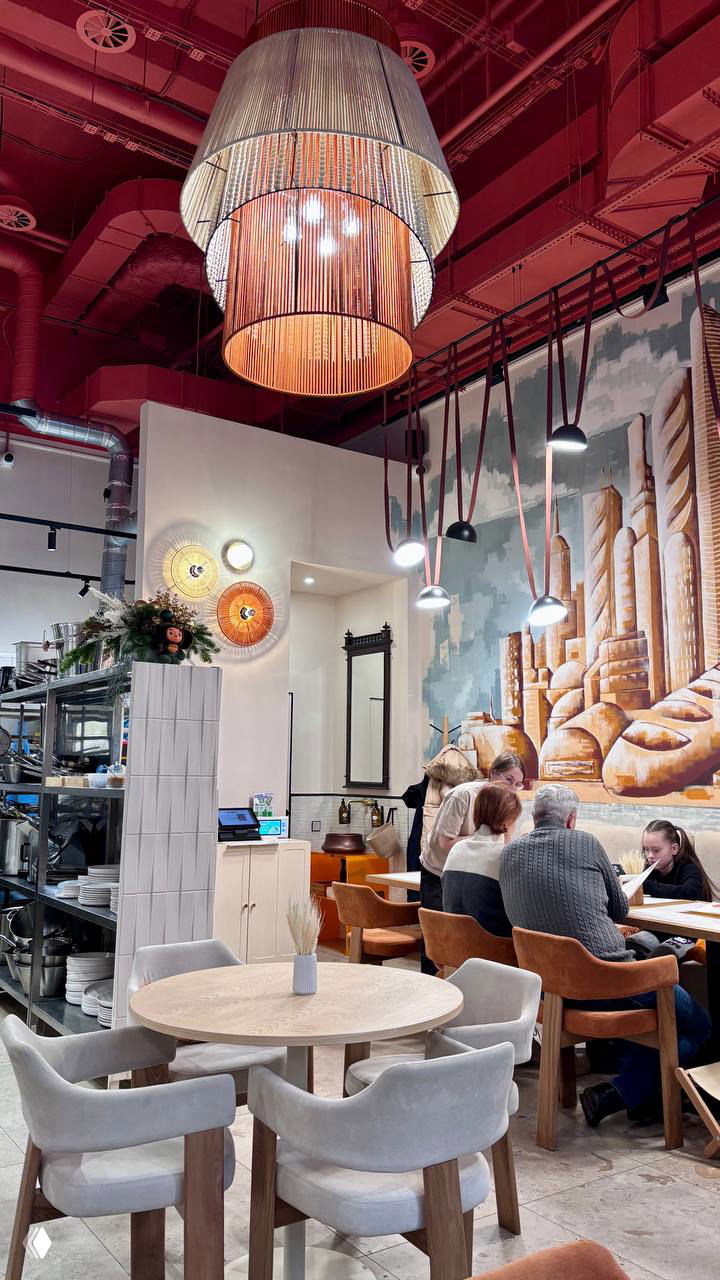 Интерьер ресторана Ухват: круглые столы, мягкие стулья, большая настенная роспись с изображением хлеба и декоративные подвесные светильники.