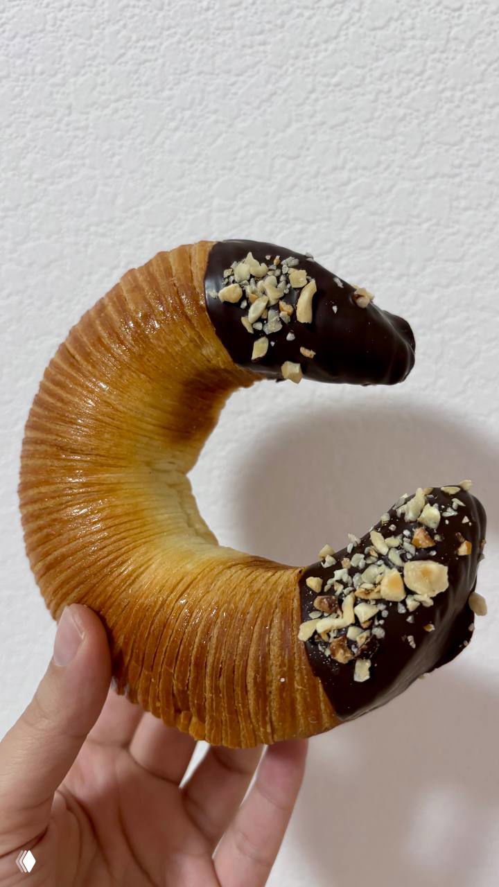 Полукруглый рогалик в руке, концы обмакнуты в шоколад и посыпаны дроблёными орехами, видно слоёное тесто и текстуру изделия.