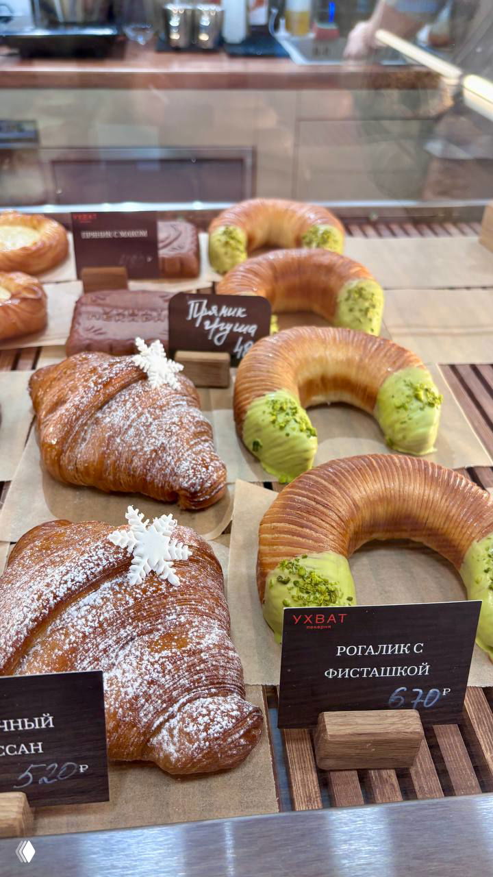Витрина пекарни с рогаликами и круассанами, рогалик с фисташковым кремом и таблички с ценами на фоне стеклянной прилавочной витрины.
