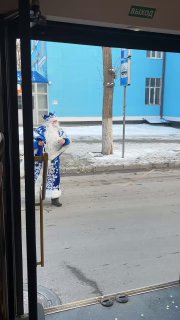 Сцена снаружи: Дед Мороз в синем костюме стоит на улице рядом с трамваем, вид через открытую дверь и городской фон за его спиной.