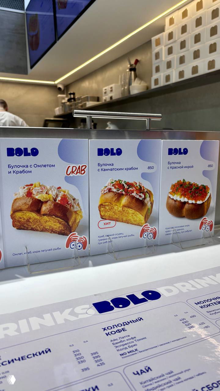 Интерьер корнера Bolo в ТРЦ: стойка заказа, меню с картинками булочек и синие стаканы для напитков на заднем плане, бариста за кассой.