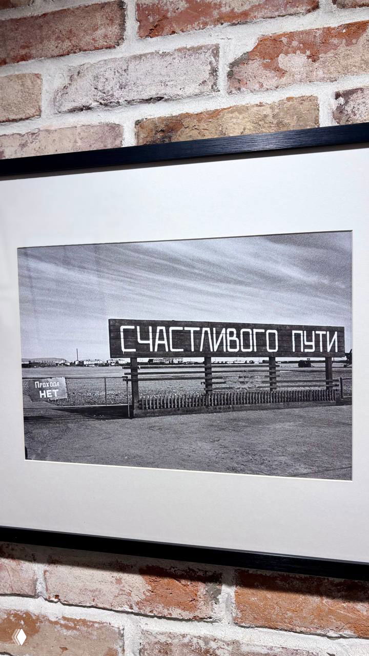 Чёрно‑белая фотография с надписью «Счастливого пути» в раме на кирпичной стене — один из экспонируемых снимков в прогулочной серии.