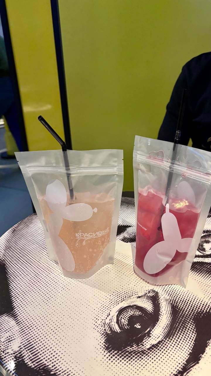 Два напитка в пластиковых пакетах с соломинками стоят на столе с принтом лица — лимонады или коктейли в фирменных пакетах бара.