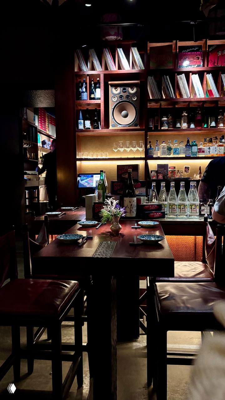 Интерьер бара в Self Edge: высокий стол, полки с бутылками и виниловыми дисками, тёплое освещение создаёт уютную атмосферу для ужина.