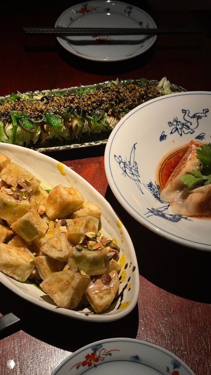 Разнообразные закуски и горячие блюда на тарелках: жареные кусочки, пельмени и длинная зелёная закуска на фоне тёплого света.