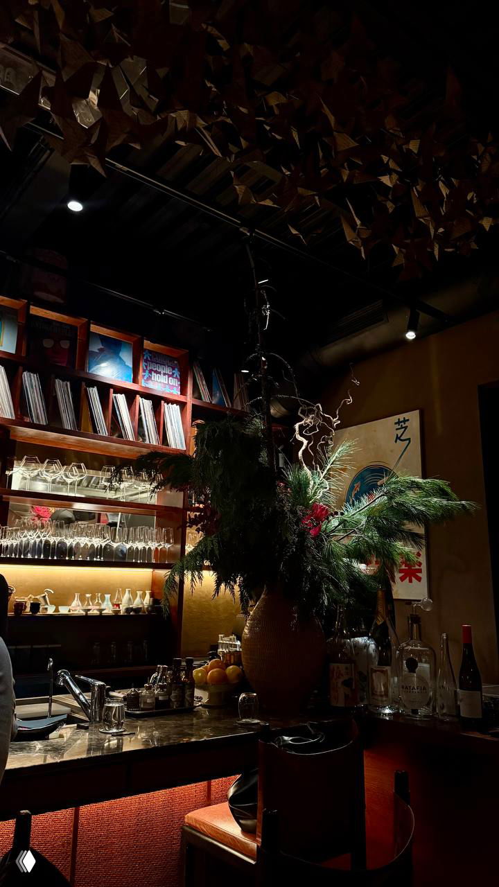 Тёмный задник бара с декоративной композицией из зелени и бутылок: атмосферное освещение и элементы декора над барной стойкой ресторана.