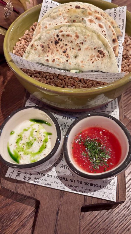 Подача: миска с тонкими лепёшками, подложенная на зерна, и две маленькие соусницы с белым и ярко‑красным соусом — контраст вкусов и текстур.