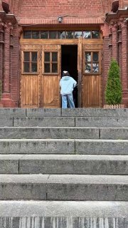 Узкий вертикальный кадр ступеней и деревянной двери собора, человек стоит перед входом, видна текстура кирпича и пара декоративных растений.