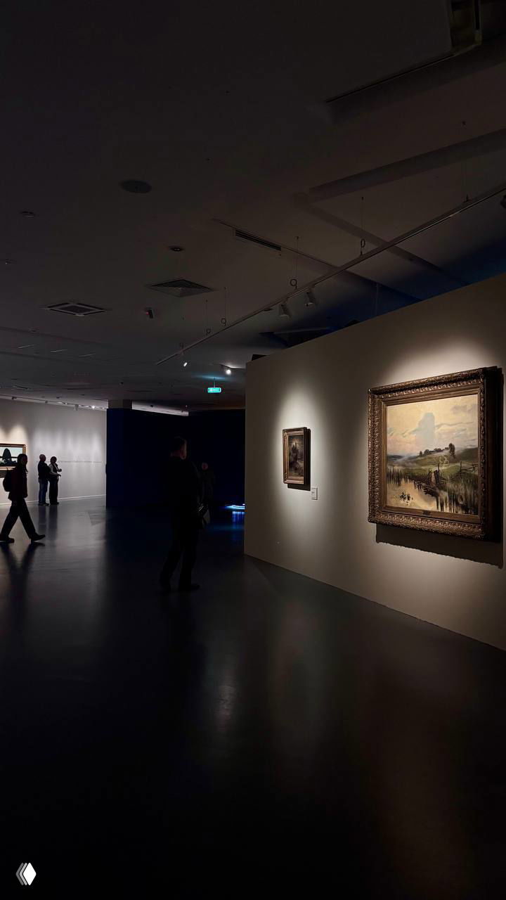 Интерьер зала музея с висящими живописными полотнами, приглушённое освещение и несколько посетителей, рассматривающих картины на стенах.