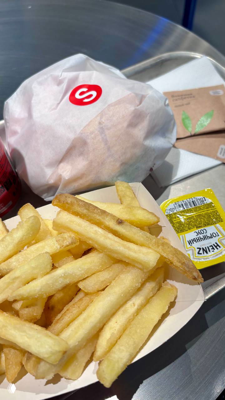 Лоток с классической картошкой фри и упакованный бургер на столе в бумажной обертке с логотипом, рядом соус Heinz — трапеза в фастфуде.