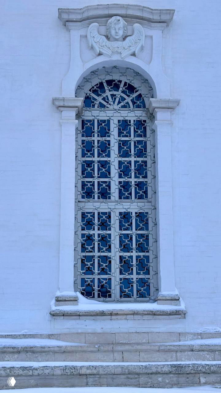 Деталь фасада и витражного окна Воскресенского собора: белая кирпичная отделка, декоративный архивольт и резной ангел над окном в снегу.