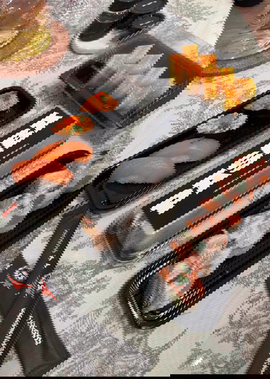 Вид сверху на контейнеры с роллами Pasiba: несколько пластиковых ланч-боксов с разными роллами, палочки, упаковка и сервировка на скатерти