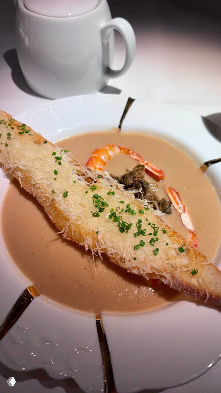 Тарелка лукового супа-пюре с крупной креветкой и длинным хрустящим багетом, покрытым тёртым сыром и мелкой зеленью в ресторанной подаче.