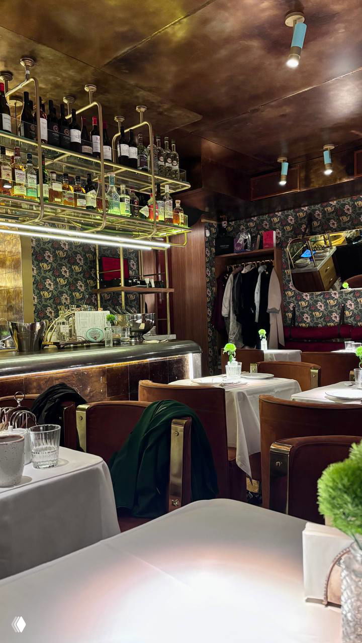 Барная зона с подвесной полкой бутылок, металлическими элементами и аккуратно накрытыми столиками в белых скатертях — вечерняя подсветка и уют.