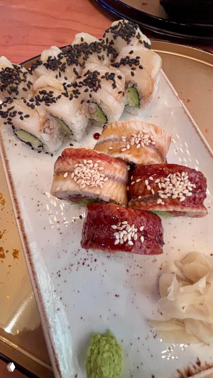 Ближний план роллов с белым и чёрным кунжутом на прямоугольной тарелке рядом с пастой васаби и маринованным имбирём — подача одного из сетов.