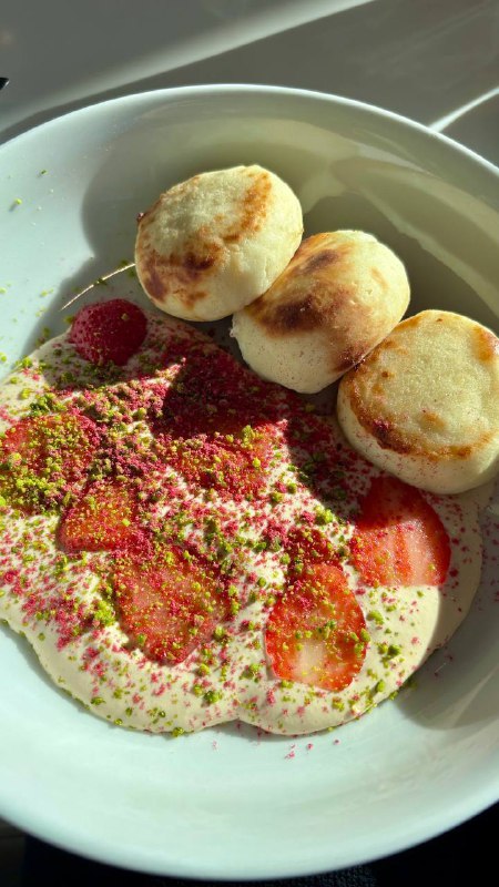 Белая миска с сырниками и кремом, рядом тонко нарезанная клубника и зелёная посыпка, крупный план десерта в кафе.