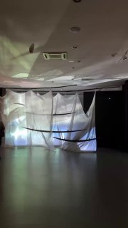 Инсталляция в выставочном зале: большие лёгкие ткани и проекция света на сценографическом пространстве, пустой зал перед показом.