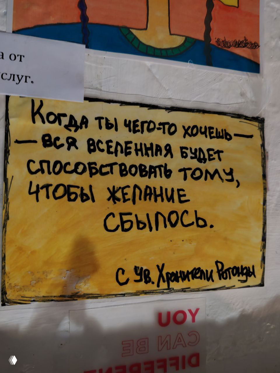 Плакат с рукописной фразой: «Когда ты чего-то хочешь — вся Вселенная будет способствовать тому, чтобы желание сбылось», заметно на стене.