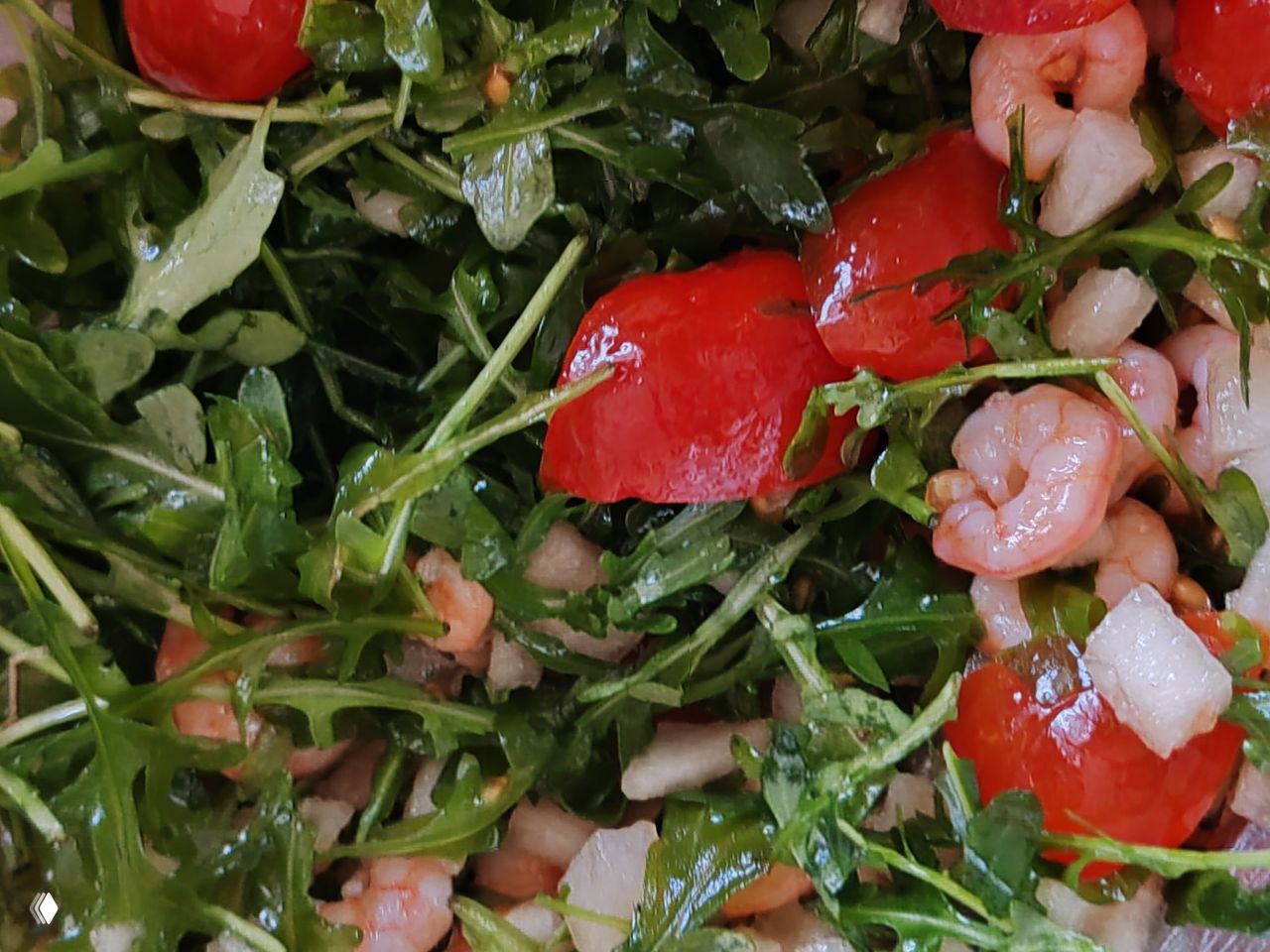 Крупный план салата из рукколы с помидорками черри, мелкими креветками и кусочками лука; видна заправка маслом.