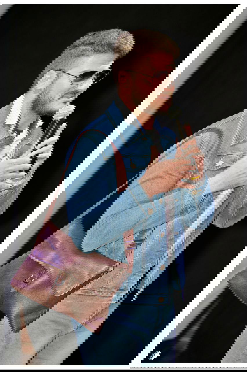 Александр в джинсовой куртке с сумкой через плечо говорит в микрофон на светлом фоне сцены, портретный кадр