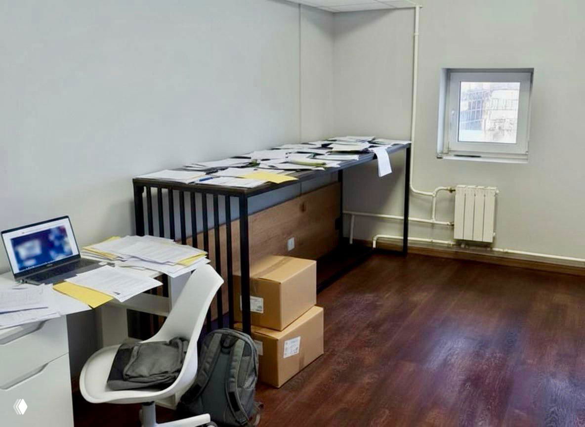 Небольшой рабочий стол с ноутбуком, стопками документов и картонными коробками — интерьер офисного склада/бэк-офиса.