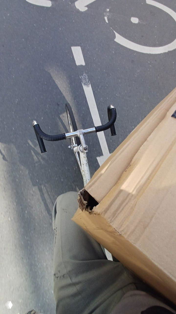 Вид с велосипеда: коробка лежит на коленях и частично торчит за рулём во время езды — демонстрация перевозки габаритной посылки верхом на велосипеде.