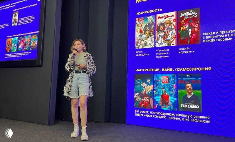 Спикер на сцене возле большого экрана с яркими слайдами о жанровости и настроении анимации; полный рост, кэжуал образ.