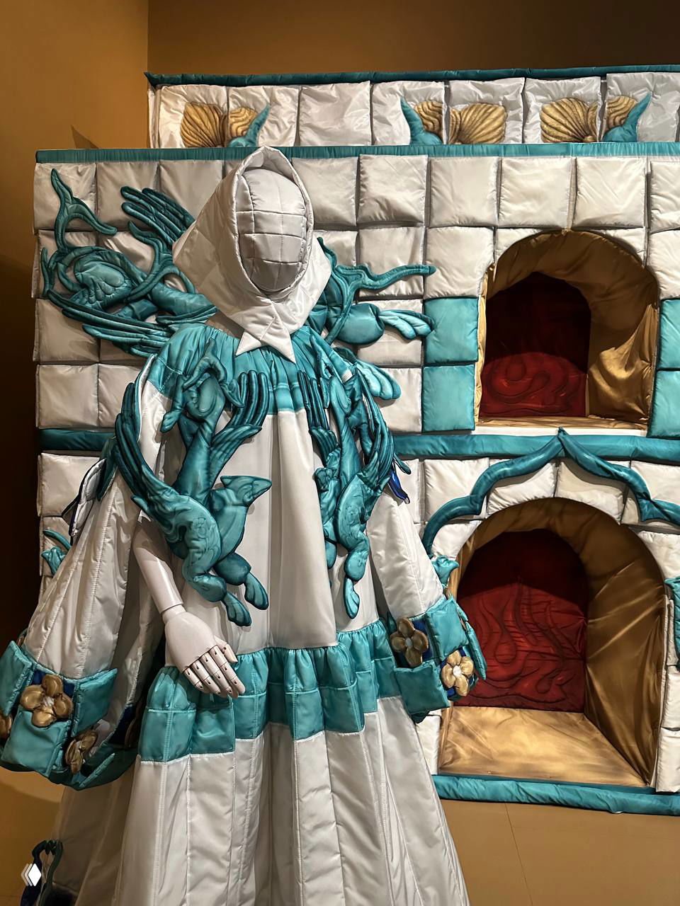Манекен в объёмном бирюзово-белом платье с бархатными и текстильными элементами на фоне мягкой текстильной инсталляции — тканевые объёмы и рельефы.