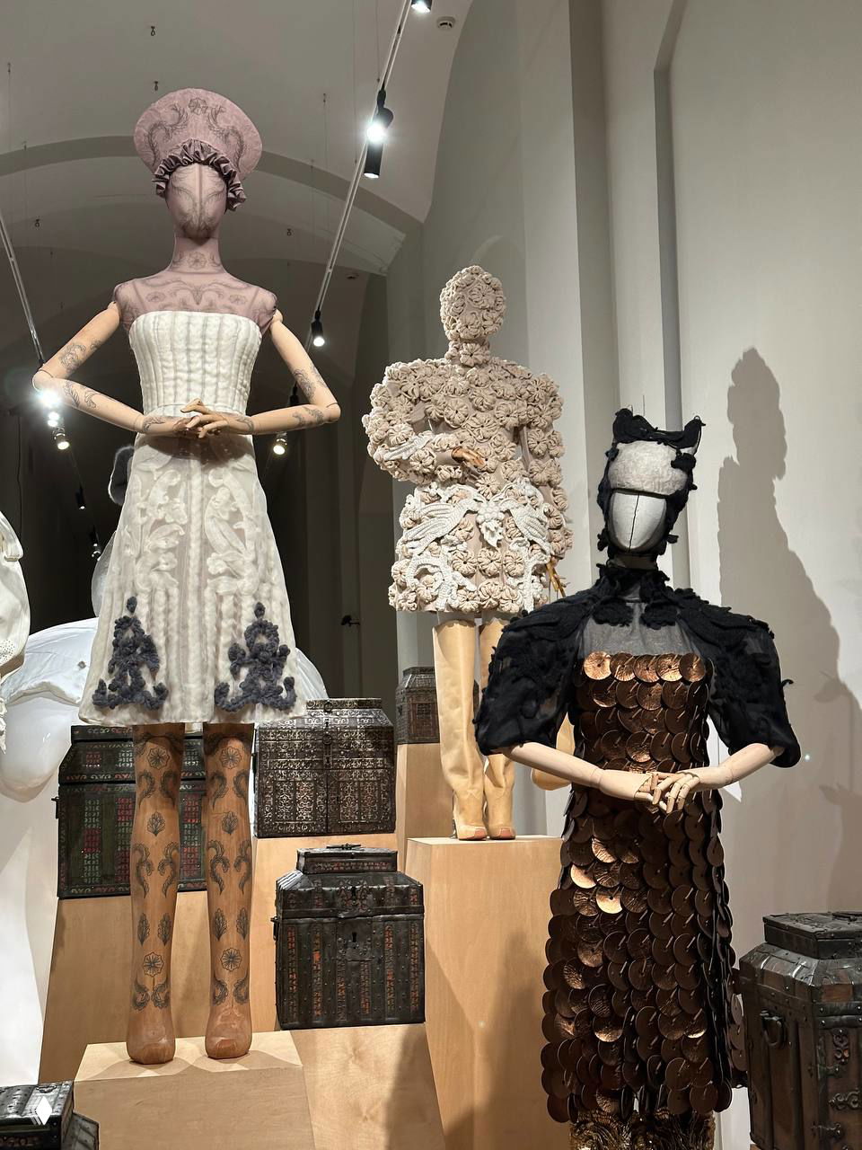 Группа манекенов в концептуальных костюмах: белые, бежевые и тёмные наряды с текстурными деталями и металлическими элементами в композиции.