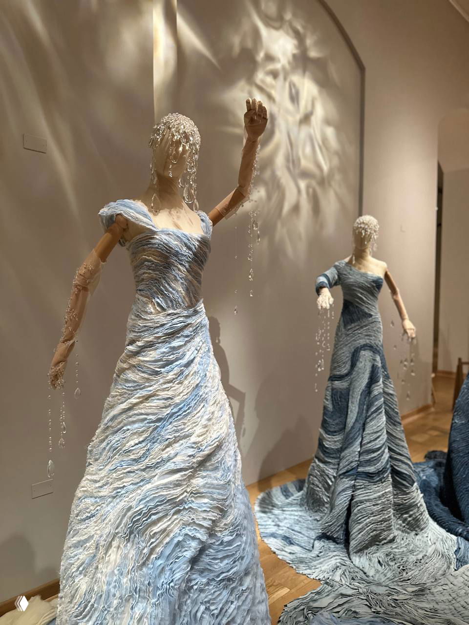 Элегантные белые платья-скульптуры с рябью текстуры и подвесными стеклянными каплями, создающими игру света и тени на стене галереи.