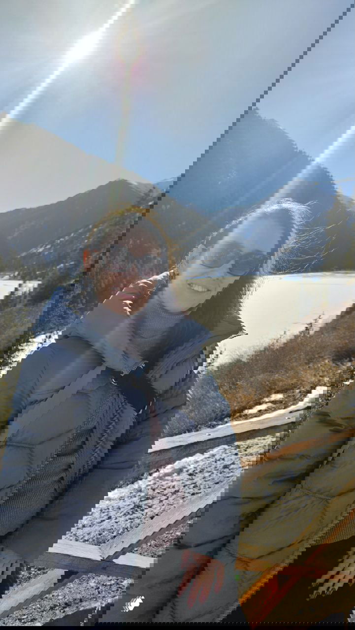 Улыбающаяся женщина в тёплой куртке на деревянном мостике у озера Кольсай, на заднем плане горы, лёд на воде и солнечный блик.