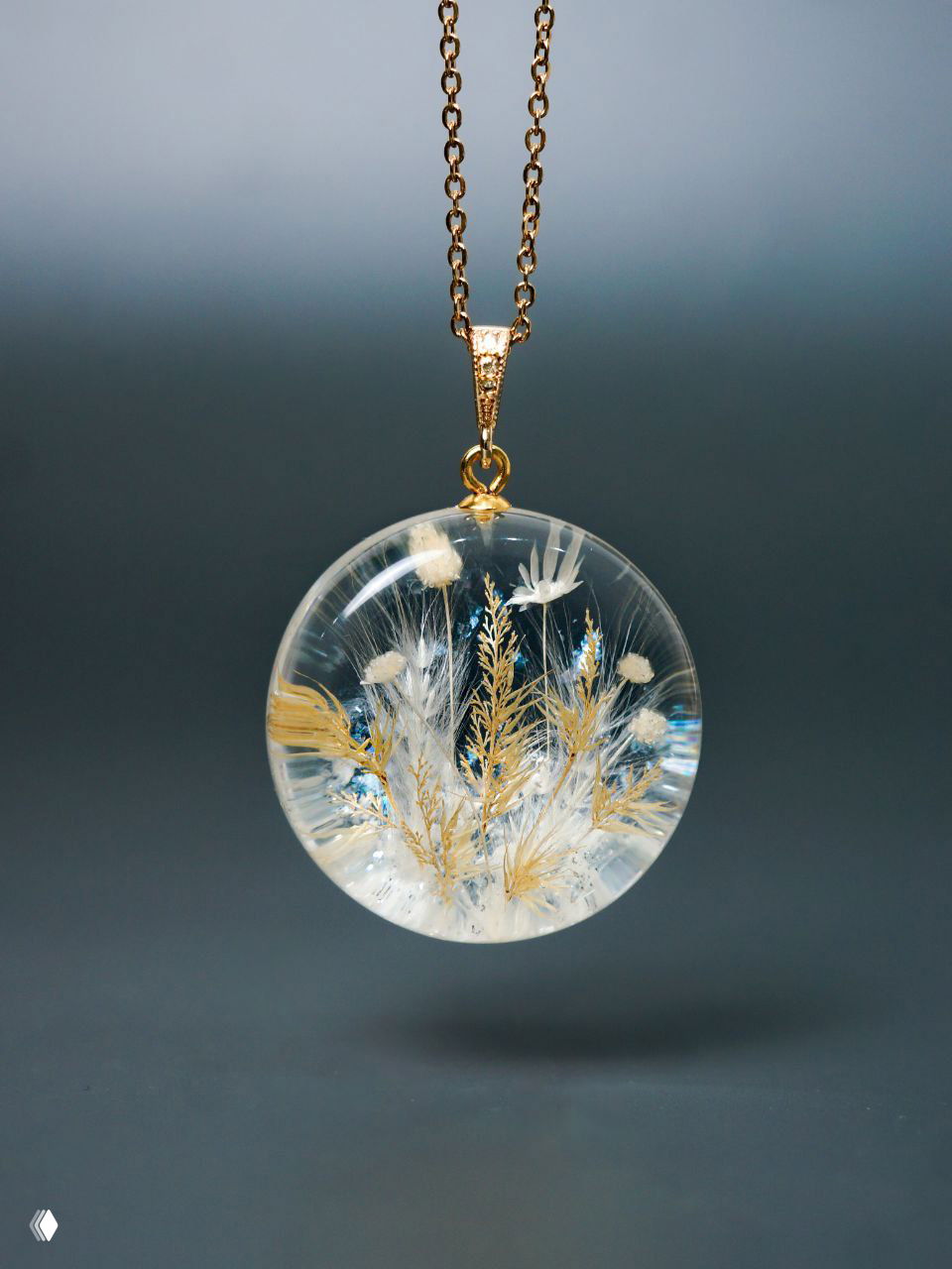 Подвеска-талисман в виде круглого прозрачного медальона с золотистыми травами и белыми акцентами, висящая на тонкой цепочке на нейтральном фоне.