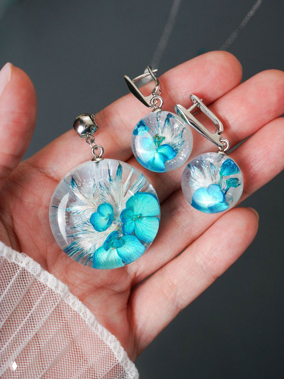 Несколько круглых кулонов разного размера с засушенными травами и мелкими соцветиями внутри смолы, показаны на ладони для сравнения масштаба и отделки.