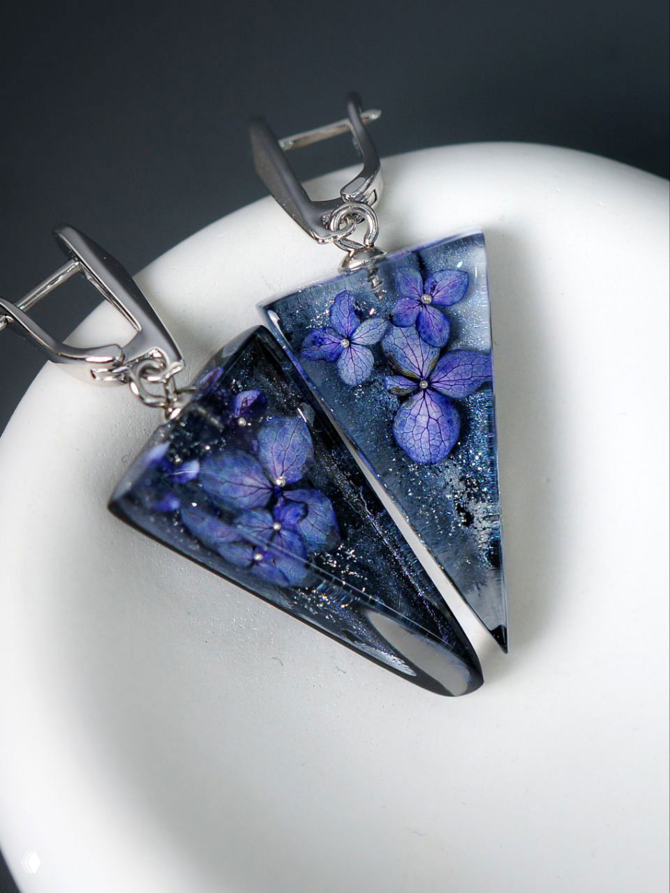 Крупный план треугольных серёжек из прозрачной смолы с застывшими синими цветками и серебристыми креплениями на светлом фоне.