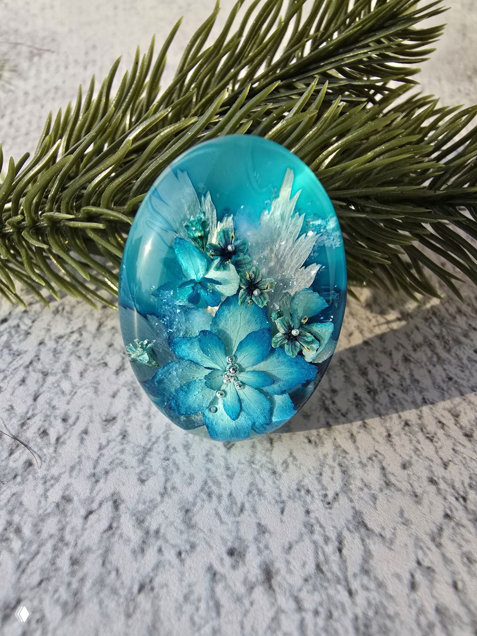 Голубой овальный кабошон с голубыми цветами и блёстками на фоне еловой ветки — завершённое украшение ручной работы.