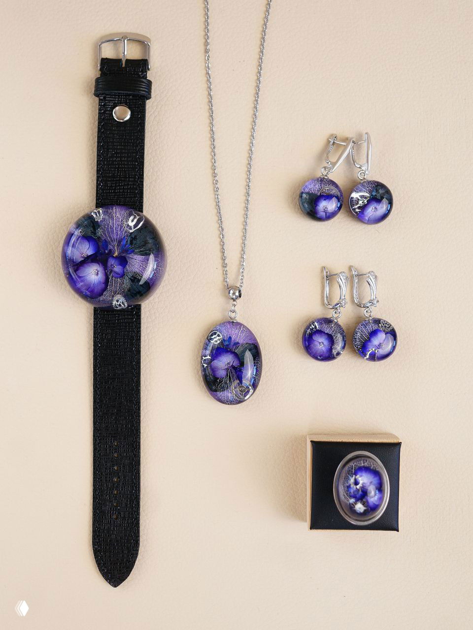Комплект украшений фиолетового оттенка: браслет, кулон и несколько пар серёг с засушенными фиолетовыми цветами в прозрачной смоле на бежевом фоне.