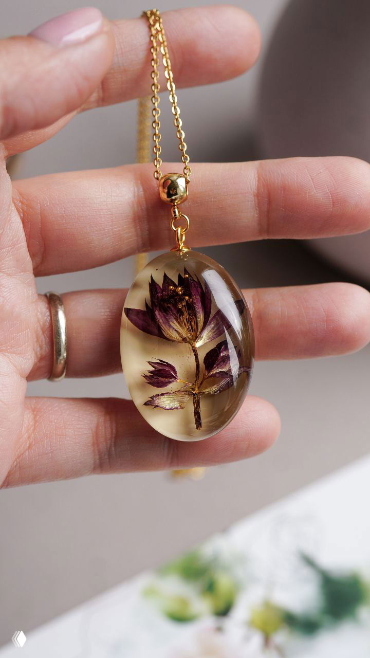 Крупный план кулона в руке: овальная прозрачная подвеска со встроенным засушенным цветком и золотистой фурнитурой, на светлом фоне.