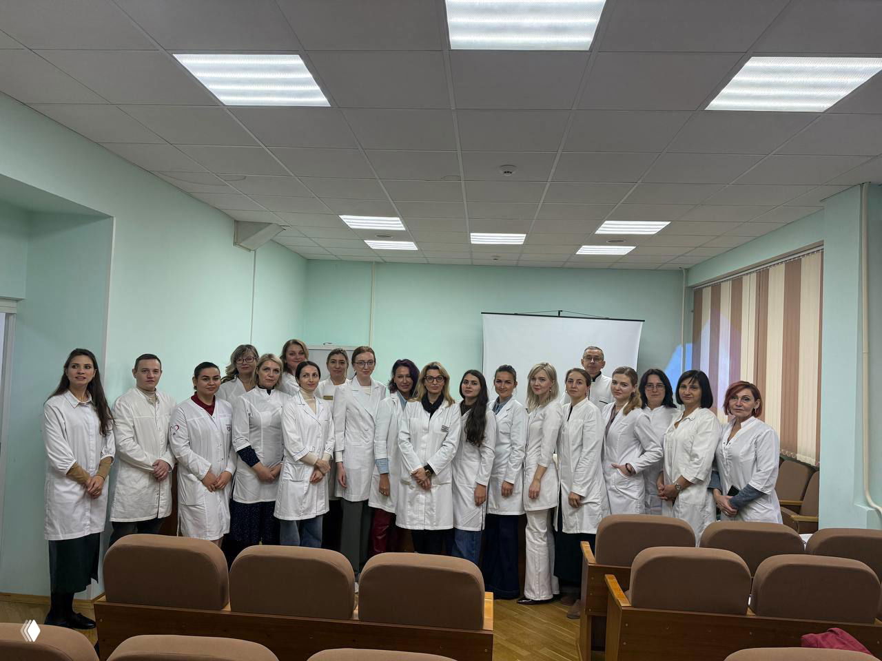 Группа врачей и слушателей в белых халатах на мастер-классе по ботулинотерапии в учебной аудитории клиники.