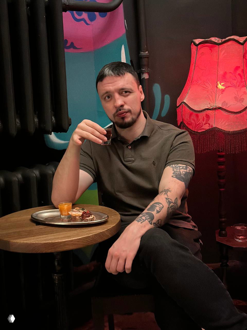 Посетитель с татуировками сидит за столиком в баре «Витя», держит рюмку; рядом торшер с тканевым абажуром и граффити на стене создают интимную сцену.