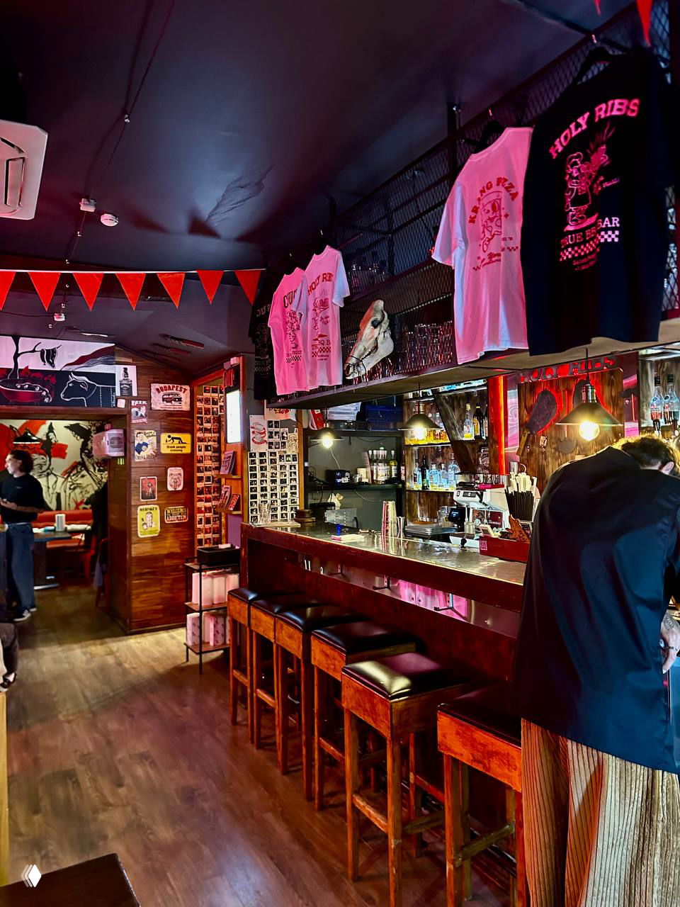 Барная стойка с табуретами, витриной и висящими футболками Holy Ribs; тёплая деревянная обстановка и плакаты на стенах.