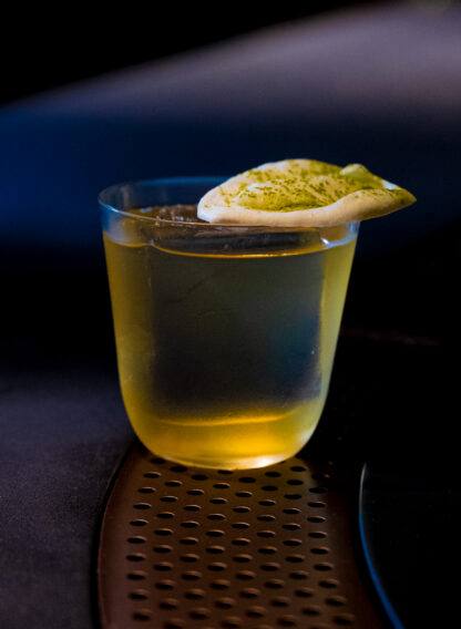 Низкий стакан с пенистой шапкой и зелёной посыпкой на поверхности; минималистичная подача авторского коктейля в приглушённой барной атмосфере.