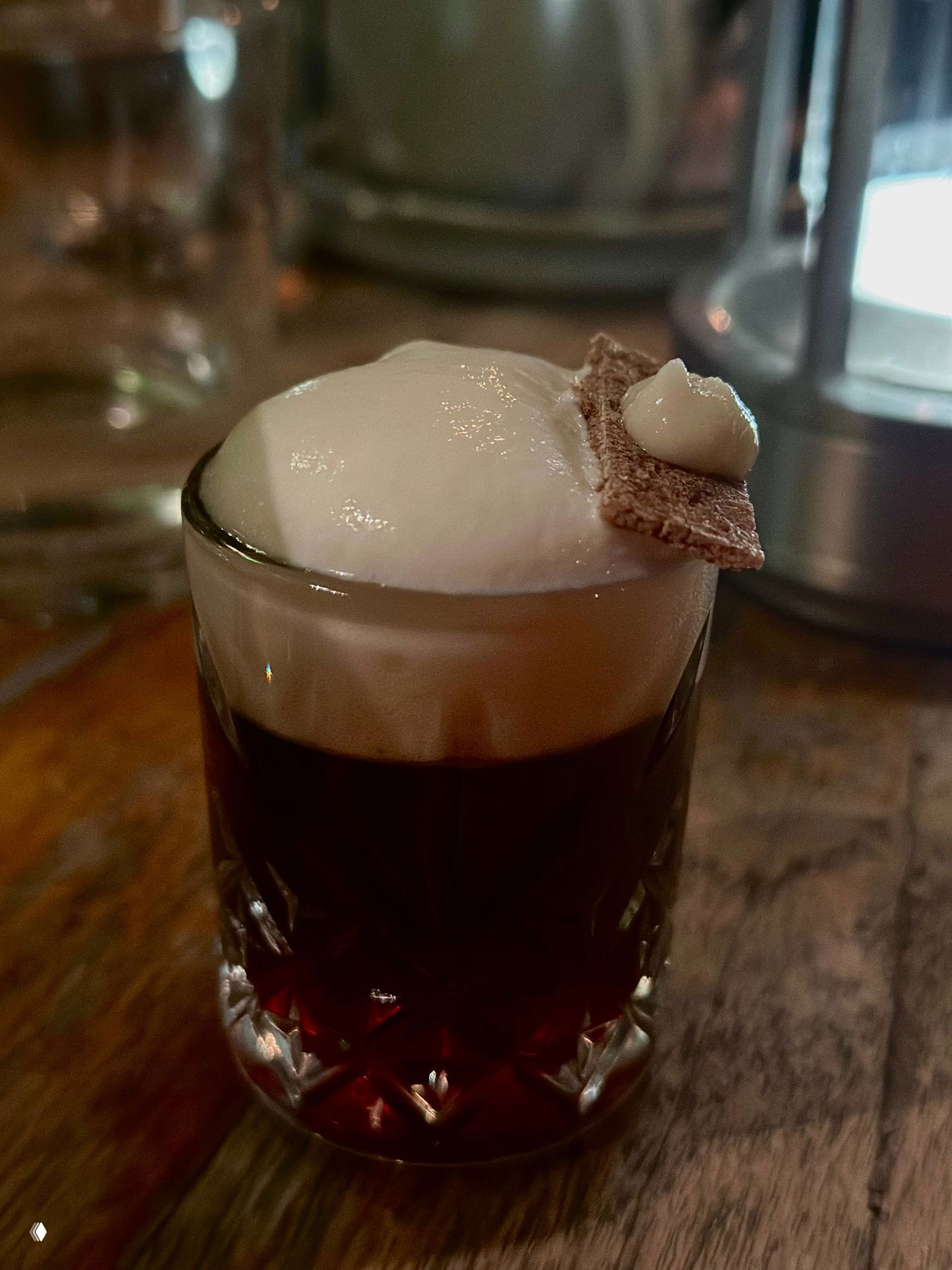 Крупный план шота «borsch» с тёмной основой и густой белой пеной, сверху гренка с кремовой точкой на деревянном столе в интерьере бара.
