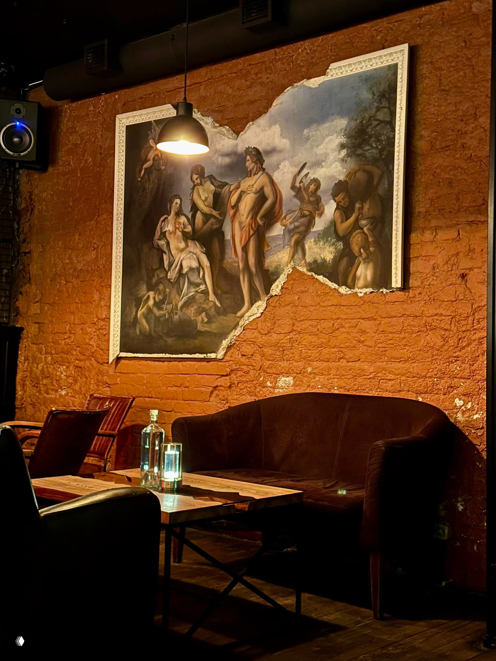 Уютная зона бара: кожаный диван, низкий стол с бутылкой и подсветкой, большая декоративная картина на частично обнажённой кирпичной стене.