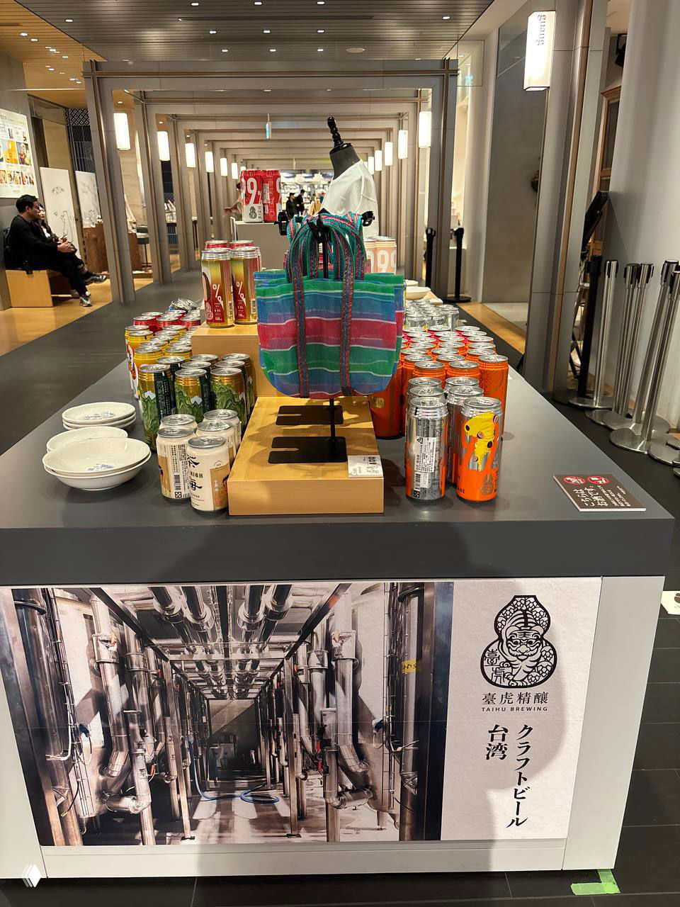 Демонстрационный стол в магазине с рядом банок тайваньских напитков, яркой полосатой сумкой и рекламным постером пивоварни на переднем плане.