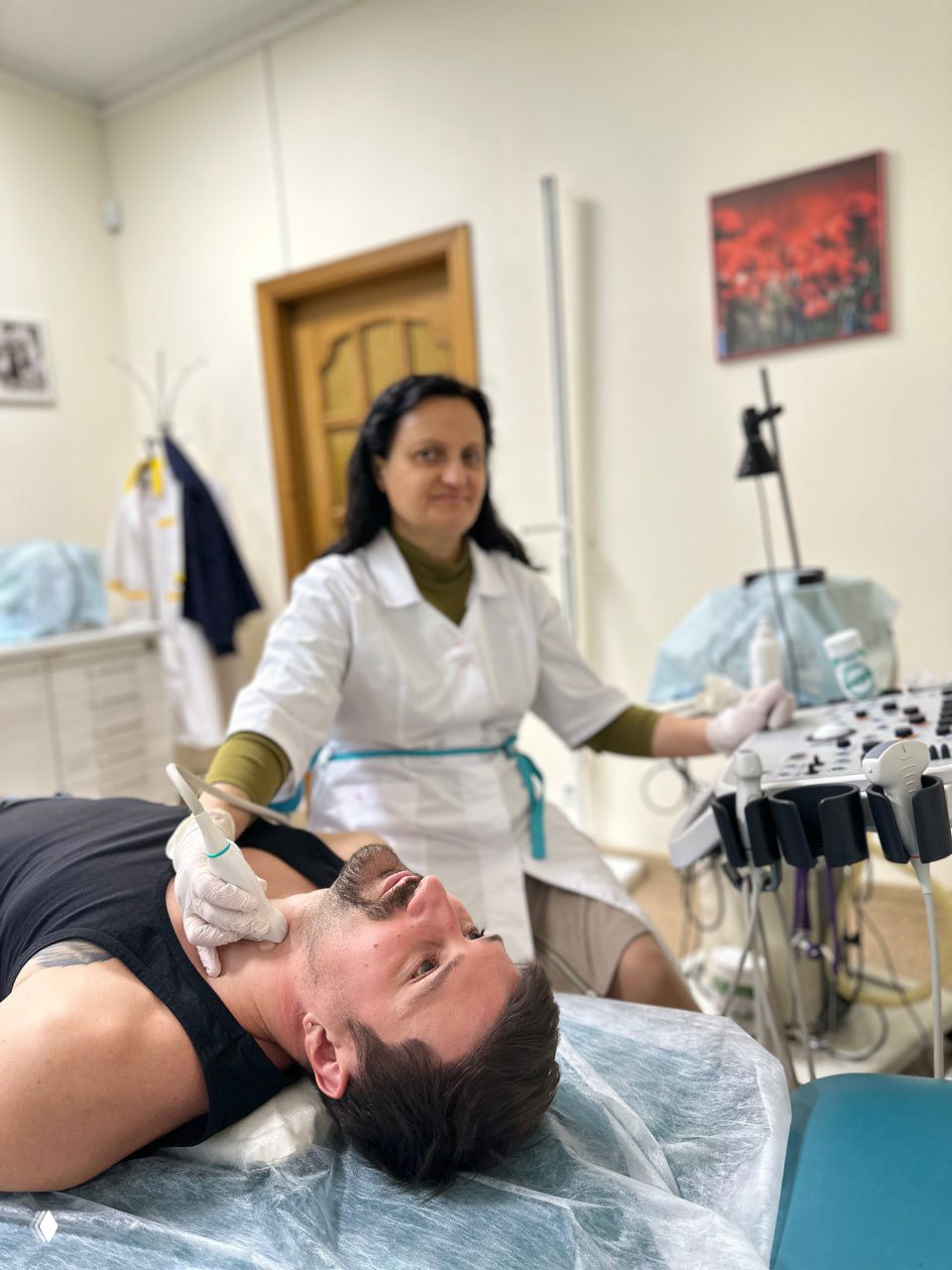 Женщина-врач в белом халате проводит обследование мужчине на кушетке; видны датчик, монитор и интерьер небольшого медицинского кабинета.