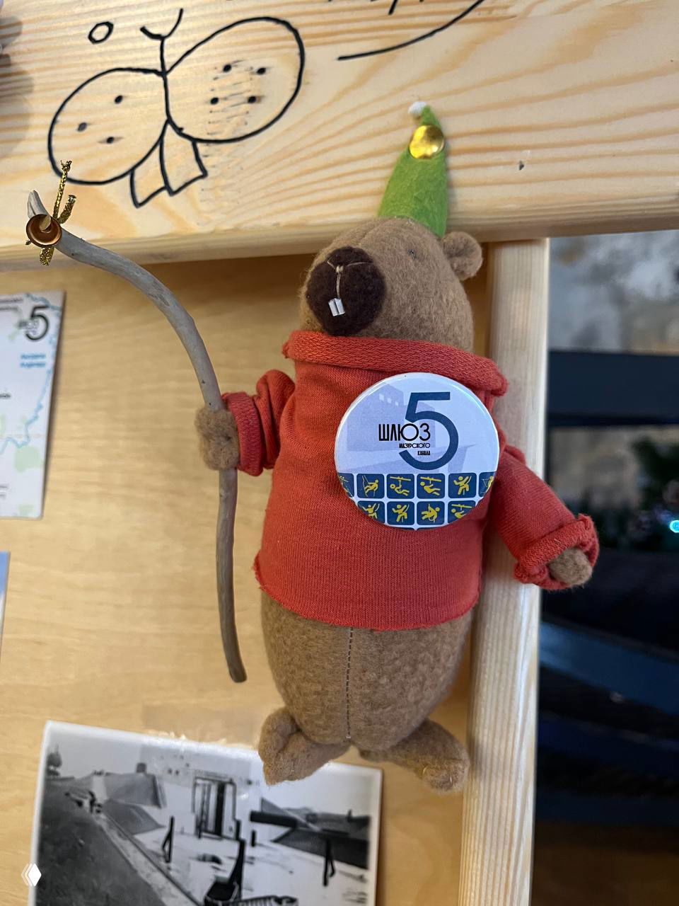 Фетровая игрушка-бобр с большой круглой кнопкой «Шлюз 5», висящая на деревянной стойке, рядом видны праздничные украшения.