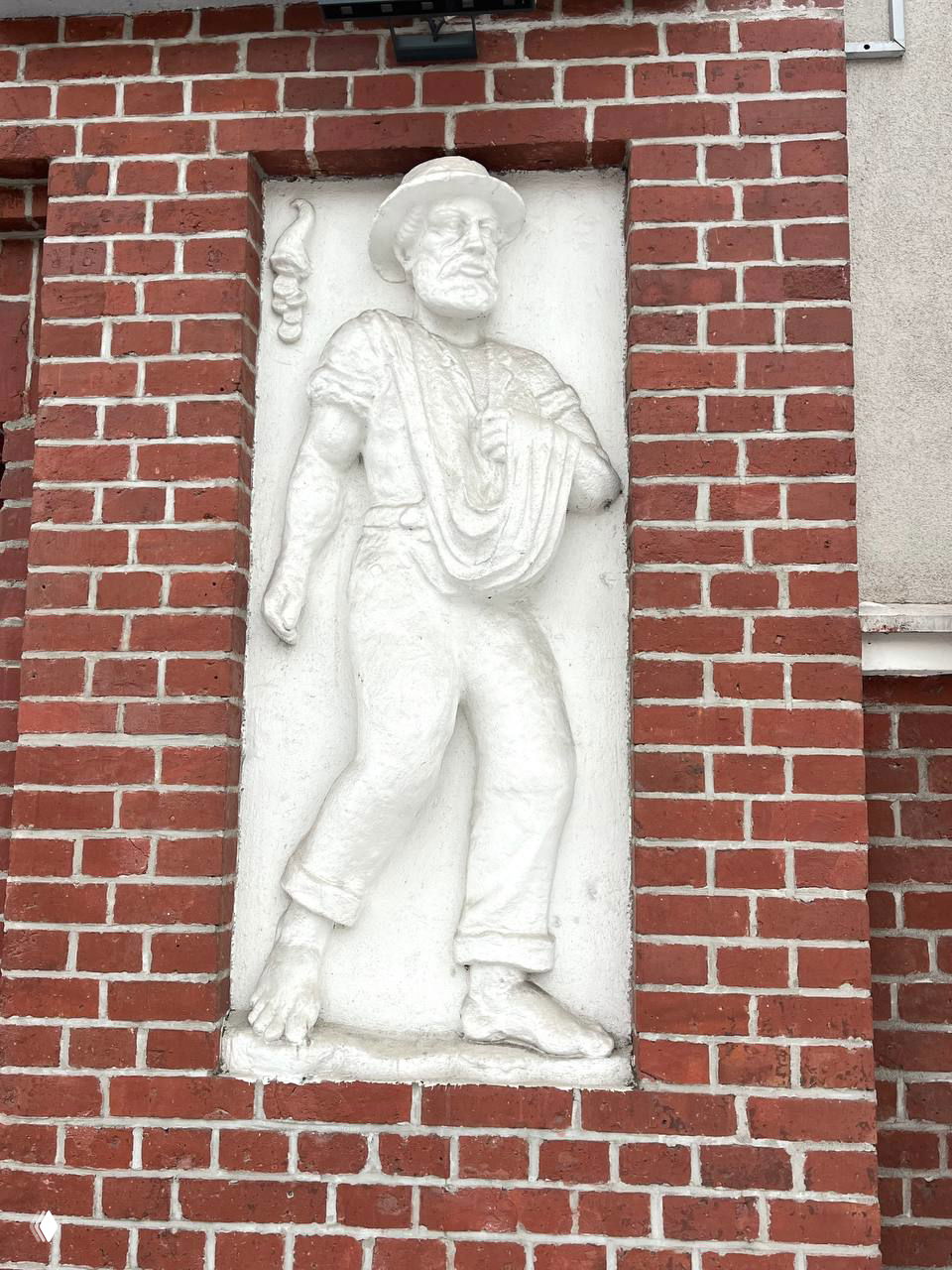 Барельеф мужского образа в нише красного кирпичного фасада; видны детали одежды, складки и рельефная фактура скульптуры в зимнем освещении.