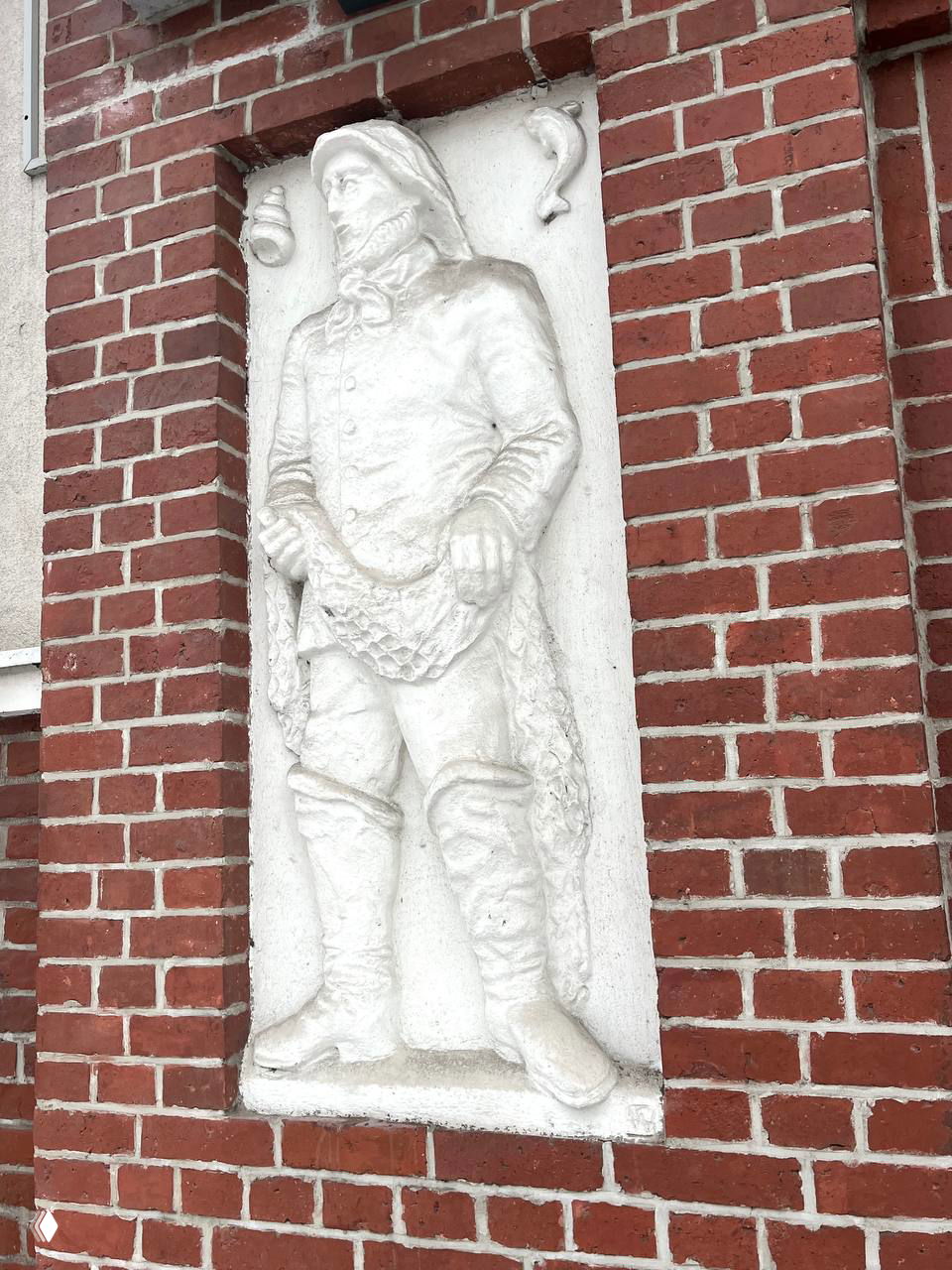 Детальный план барельефа рыбака в белой штукатурке на фоне кирпича; видны рыба, сеть и атрибуты морской профессии в нише.