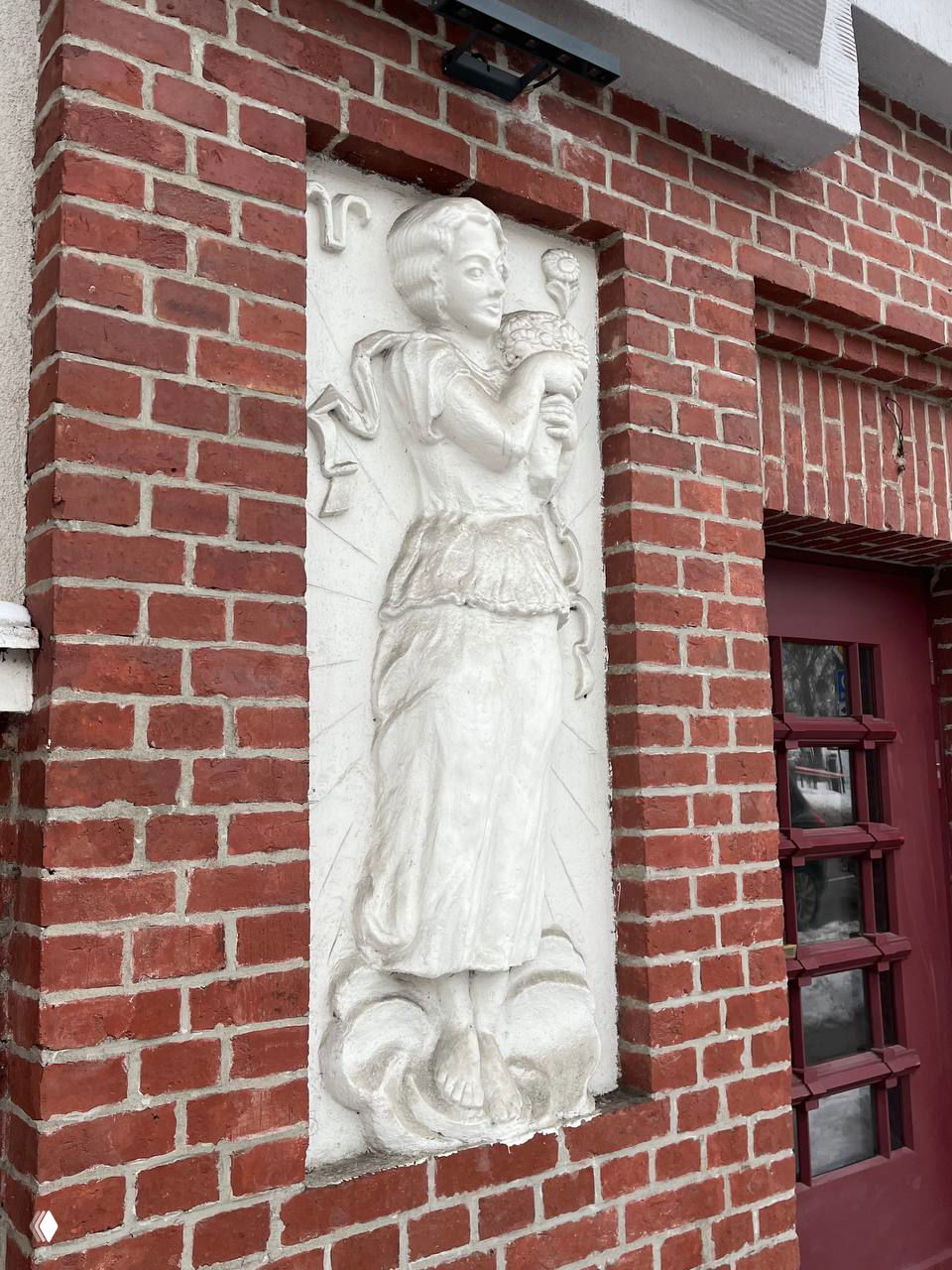 Женская фигура в нише фасада держит цветок; рядом заметен зодиакальный знак, скульптура выполнена в белой штукатурке на красном кирпиче.